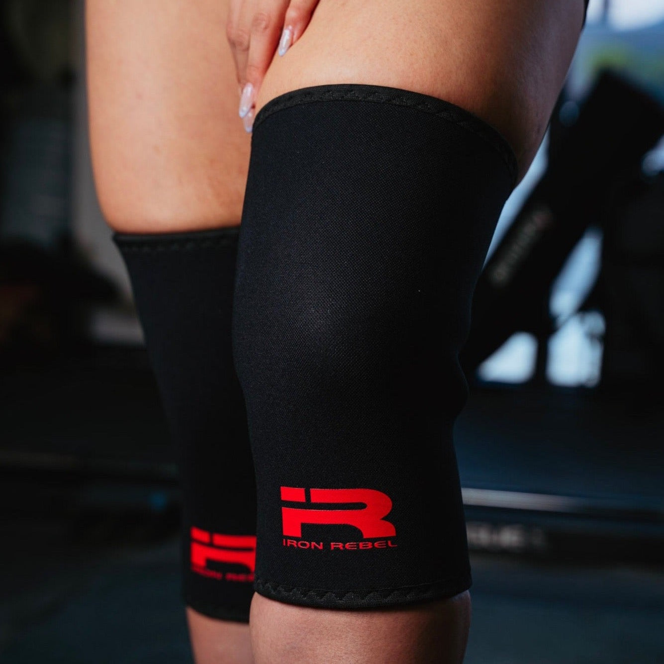 Close-up of a female lifter wearing black Iron Rebel PR Cone Knee Sleeves with a red Iron Rebel logo, showing a snug fit over the knees. Provides firm neoprene compression and joint support for powerlifting, squats, and heavy weightlifting workouts.