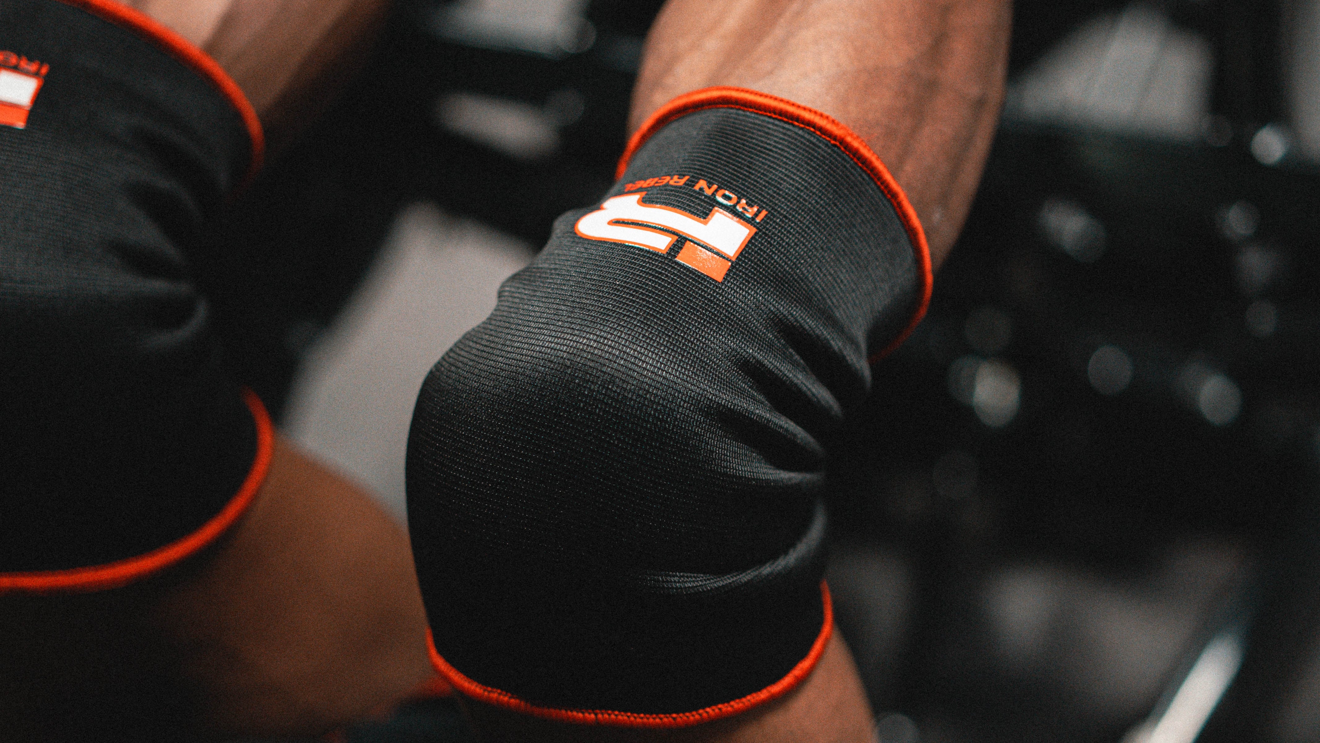 Close-up of a person lying on a gym bench pressing, wearing black knee sleeves with orange trim and a visible logo, along with a black shirt in a gym setting.